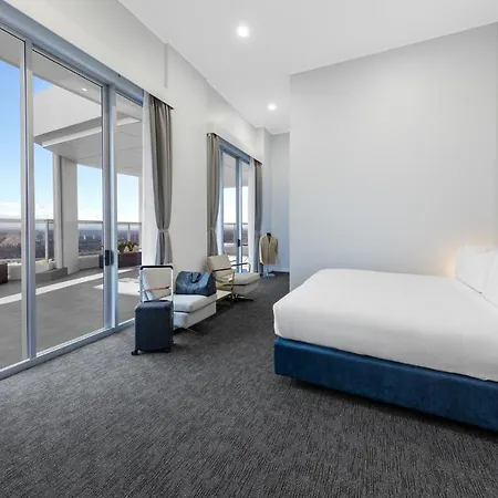 Meriton Suites Bondi Junction Sydney Exterior photo The photo shows a modern and spacious hotel room. It features a large bed with a blue headboard, a flat-screen TV mounted on the wall, and a cozy seating area with a chair and small table. The room has large windows that offer natural light and a vie
