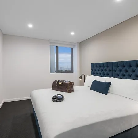Meriton Suites Bondi Junction Sydney Exterior photo The photo shows a modern bedroom featuring a large bed with a tufted blue headboard and white bedding. There is a pillow and a small dark-colored bag on the bed. A pair of headphones is also placed on the bed. On the wall opposite the bed, there is a