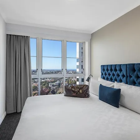 Meriton Suites Bondi Junction Sydney Exterior photo The photo shows a modern hotel room featuring a large bed with a tufted blue headboard. Beside the bed, there is a stylish bag placed on the bed. The room has large windows that provide a view of the outside, allowing natural light to fill the space.