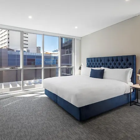 Meriton Suites Bondi Junction Sydney Exterior photo The image shows a modern hotel room with a large, comfortable bed featuring a blue upholstered headboard. The room has large windows that allow natural light to enter, providing a good view of the surrounding buildings. There is a small bedside table