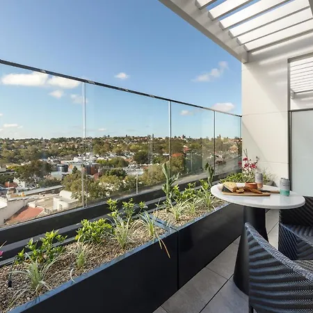 Meriton Suites Bondi Junction Sydney Exterior photo The photo shows a modern balcony with a glass railing, enhancing the view of the surrounding area, which includes various rooftops and greenery in the distance. There is a small round table set with a decorative floral arrangement and some items, lik