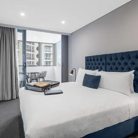 Meriton Suites Bondi Junction Sydney Exterior photo The photo shows a modern hotel room featuring a large bed with a tufted blue headboard. There are minimalistic bedside tables with lamps on either side of the bed. A flat-screen TV is mounted on the wall across from the bed. To one side, there are fl