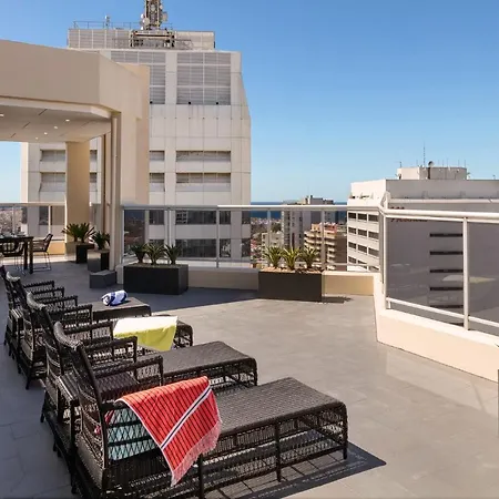 Meriton Suites Bondi Junction Sydney Exterior photo The photo shows a rooftop terrace with a modern design. There are several outdoor seating arrangements featuring black wicker lounge chairs. Some of the chairs have colorful blankets draped over them. The terrace is surrounded by a railing and has po