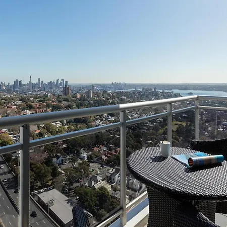 Meriton Suites Bondi Junction Sydney Exterior photo The photo shows a balcony with a view overlooking a cityscape and a body of water in the distance. There is a round table with a woven design and two chairs, one of which has a mug on it. On the table, there’s also a rolled-up magazine or document. T