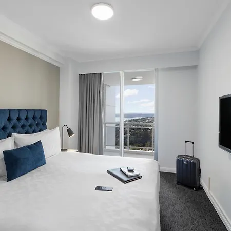 Meriton Suites Bondi Junction Sydney Exterior photo The photo shows a modern hotel room featuring a neatly made bed with a blue tufted headboard and a white comforter. A decorative blue pillow is placed on the bed. There's a bedside lamp and a small table with a phone and a notebook on it. A suitcase 