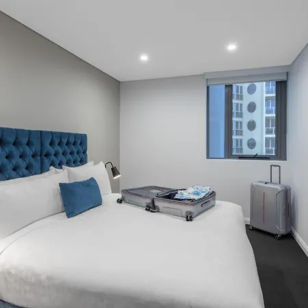 Meriton Suites Bondi Junction Sydney Exterior photo The photo shows a modern hotel room featuring a neatly made bed with a blue tufted headboard. There is a small blue decorative pillow on the bed, and the bedding is white. Next to the bed, there is a nightstand with a lamp. A suitcase is partially op