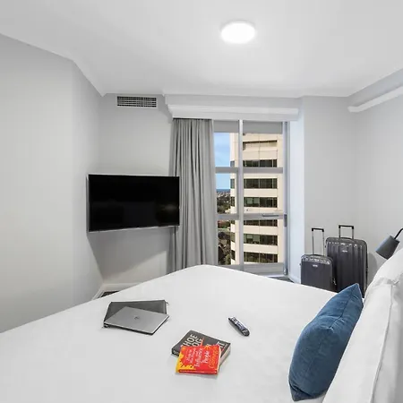 Meriton Suites Bondi Junction Sydney Exterior photo The photo shows a modern hotel room. It features a bed with white linens and blue pillows. There is a flat-screen TV mounted on the wall opposite the bed. Next to the bed, a small table holds a laptop and some books. Large windows with curtains allow