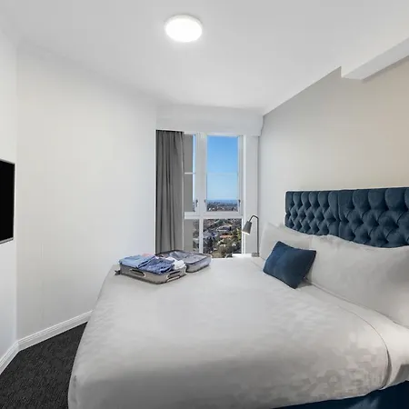 Meriton Suites Bondi Junction Sydney Exterior photo The photo shows a modern hotel room with a queen-sized bed featuring a tufted blue headboard. The bed is made with white linens and has a blue decorative pillow. On one side of the room, there's a flat-screen TV mounted on the wall. Large windows pro