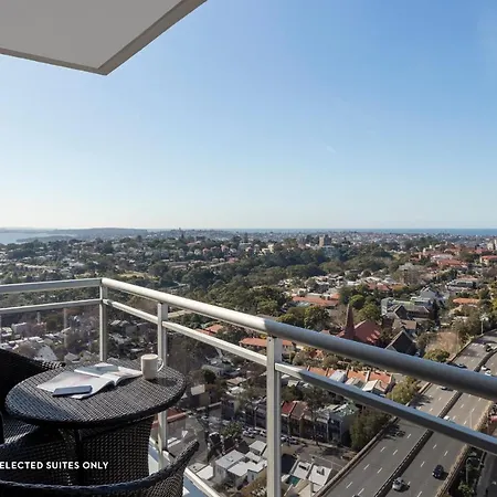 Meriton Suites Bondi Junction Sydney Exterior photo The photo shows a balcony with a view over a cityscape that includes buildings and greenery, extending towards the horizon where the sea is visible. The balcony features a small round table and two black chairs, suggesting a space for relaxation or d