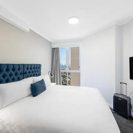 Meriton Suites Bondi Junction Sydney Exterior photo The photo shows a modern hotel room with a cozy atmosphere. It features a large bed with a tufted, blue headboard and white bedding. There is a decorative blue pillow on the bed. A small luggage suitcase is placed next to the bed. On the opposite wal