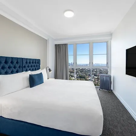Meriton Suites Bondi Junction Sydney Exterior photo The photo shows a modern hotel room featuring a large bed with a white duvet and decorative pillows. The headboard is upholstered in a deep blue fabric. There is a bedside table with a lamp next to the bed. On the opposite wall, there is a flat-scree
